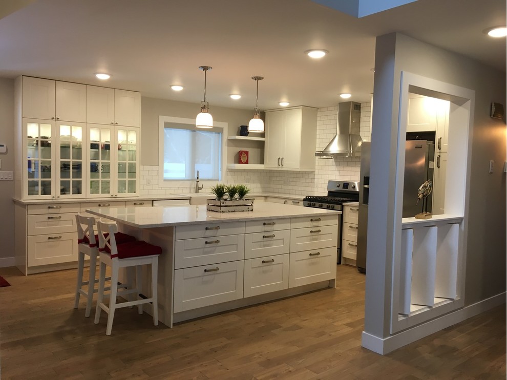 Large country l-shaped kitchen in Other with a belfast sink, white splashback, metro tiled splashback, stainless steel appliances, medium hardwood flooring, shaker cabinets, white cabinets, composite countertops and an island.