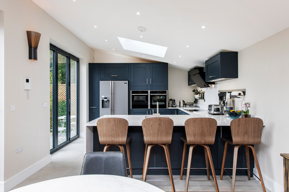 Photo of a contemporary u-shaped kitchen/diner in Hertfordshire with a submerged sink, shaker cabinets, black cabinets, white splashback, black appliances, a breakfast bar, beige floors and white worktops.