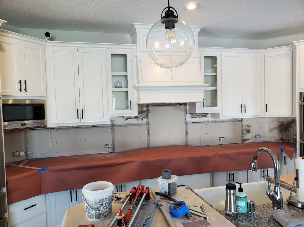 Bild på ett mellanstort funkis grå linjärt grått kök med öppen planlösning, med en rustik diskho, skåp i shakerstil, vita skåp, granitbänkskiva, vitt stänkskydd, stänkskydd i tunnelbanekakel, rostfria vitvaror och en köksö