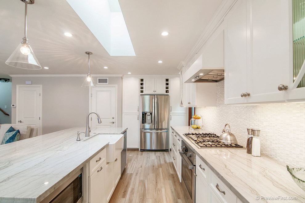 Inspiration for a mid-sized farmhouse l-shaped light wood floor open concept kitchen remodel in Los Angeles with a farmhouse sink, recessed-panel cabinets, white cabinets, marble countertops, white backsplash, mosaic tile backsplash, stainless steel appliances and an island