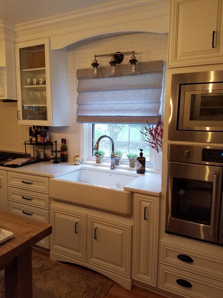 Mid-sized elegant single-wall medium tone wood floor eat-in kitchen photo in Indianapolis with a farmhouse sink, raised-panel cabinets, white cabinets, quartz countertops, white backsplash, subway tile backsplash, stainless steel appliances, an island and white countertops