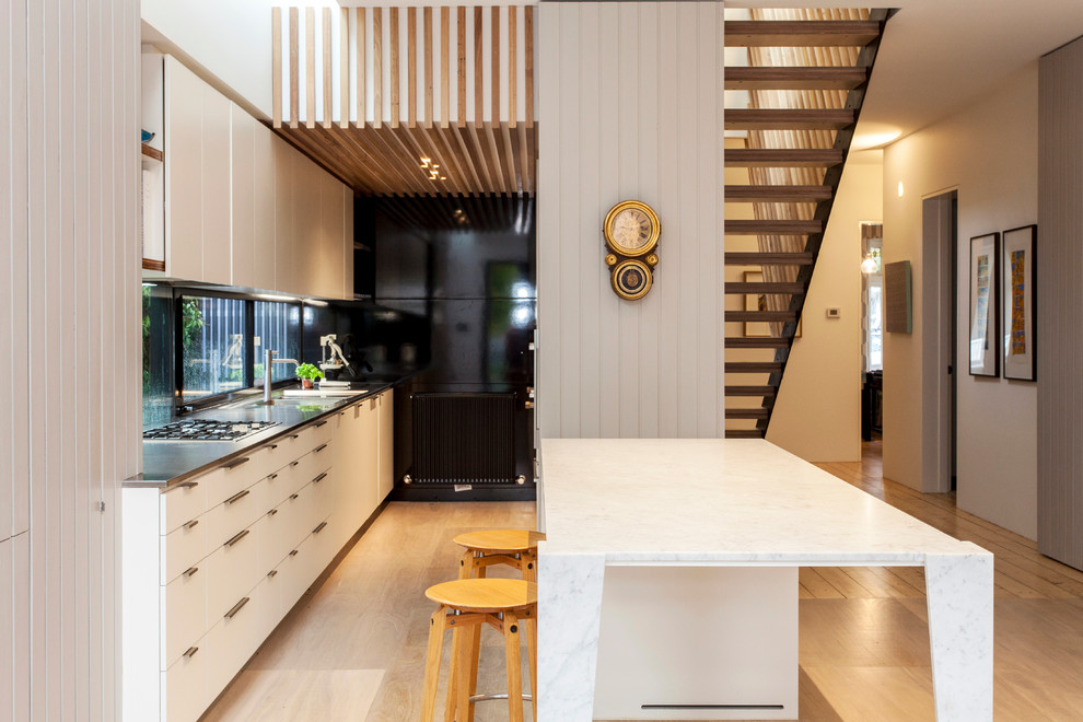 Example of a trendy galley plywood floor kitchen design in Melbourne with flat-panel cabinets, white cabinets, stainless steel countertops and an island