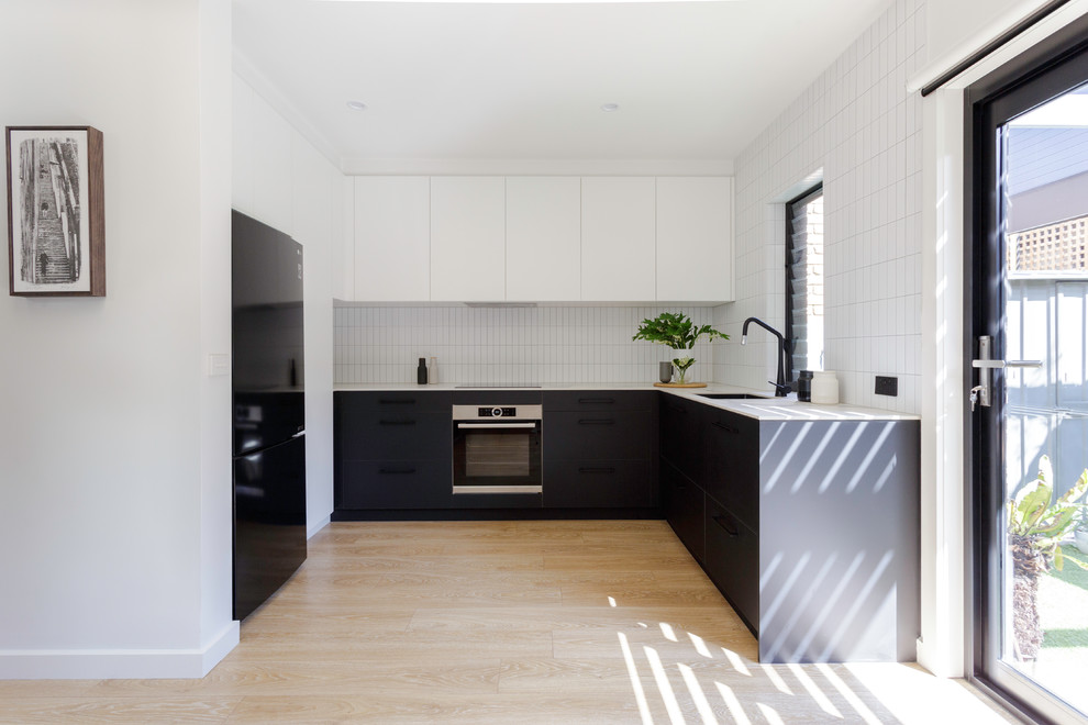 Bild på ett mellanstort funkis vit vitt u-kök, med svarta skåp, bänkskiva i rostfritt stål, vitt stänkskydd, svarta vitvaror, ljust trägolv, en undermonterad diskho, släta luckor och beiget golv