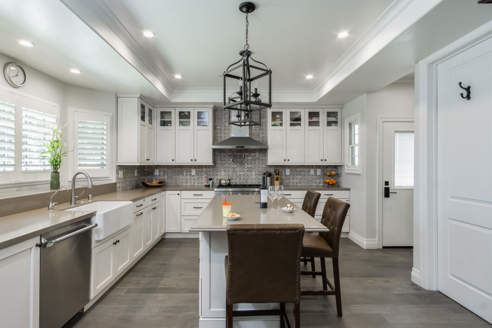This is an example of a large traditional l-shaped kitchen in Los Angeles with a belfast sink, recessed-panel cabinets, white cabinets, composite countertops, mosaic tiled splashback, stainless steel appliances, porcelain flooring, an island, beige worktops, grey splashback and grey floors.