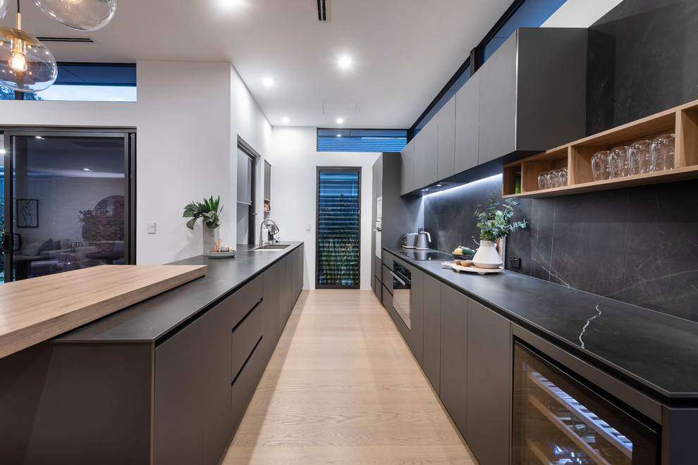 На фото: параллельная кухня в современном стиле с плоскими фасадами, серыми фасадами, серым фартуком, черной техникой, светлым паркетным полом, бежевым полом и серой столешницей с