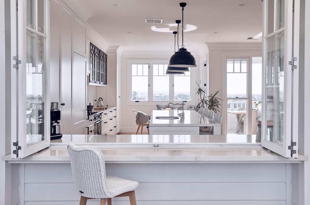 Inspiration for a large beach style galley kitchen in Sydney with a submerged sink, recessed-panel cabinets, white cabinets, white splashback, integrated appliances, medium hardwood flooring, an island, brown floors and white worktops.