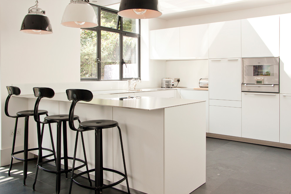 This is an example of a small contemporary u-shaped kitchen/diner in London with a submerged sink, flat-panel cabinets, white cabinets, stainless steel appliances, a breakfast bar, black floors, white splashback and white worktops.