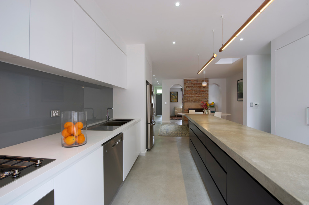 Inredning av ett modernt linjärt kök, med en dubbel diskho, släta luckor, vita skåp, bänkskiva i betong, grått stänkskydd, rostfria vitvaror, betonggolv och en köksö