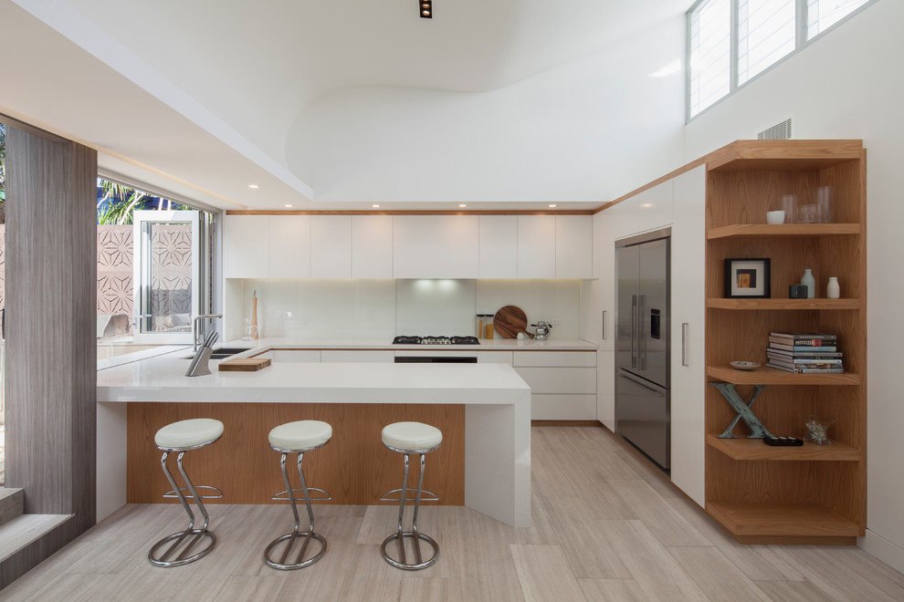 Foto på ett mellanstort funkis kök, med vita skåp, bänkskiva i kvartsit, vitt stänkskydd, glaspanel som stänkskydd, rostfria vitvaror, travertin golv, en undermonterad diskho, släta luckor och en halv köksö