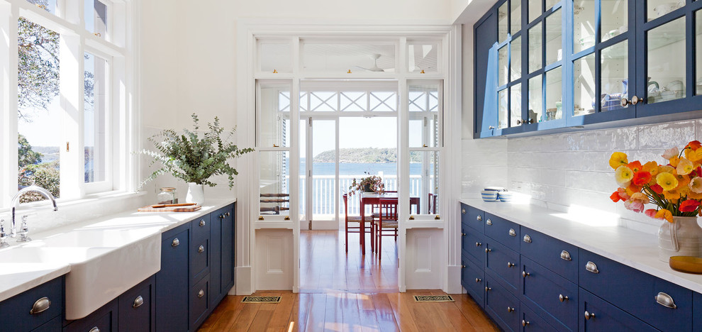 Cette photo montre une grande cuisine parallèle bord de mer avec un évier 2 bacs, des portes de placard bleues, plan de travail en marbre, une crédence blanche, une crédence en céramique et parquet foncé.