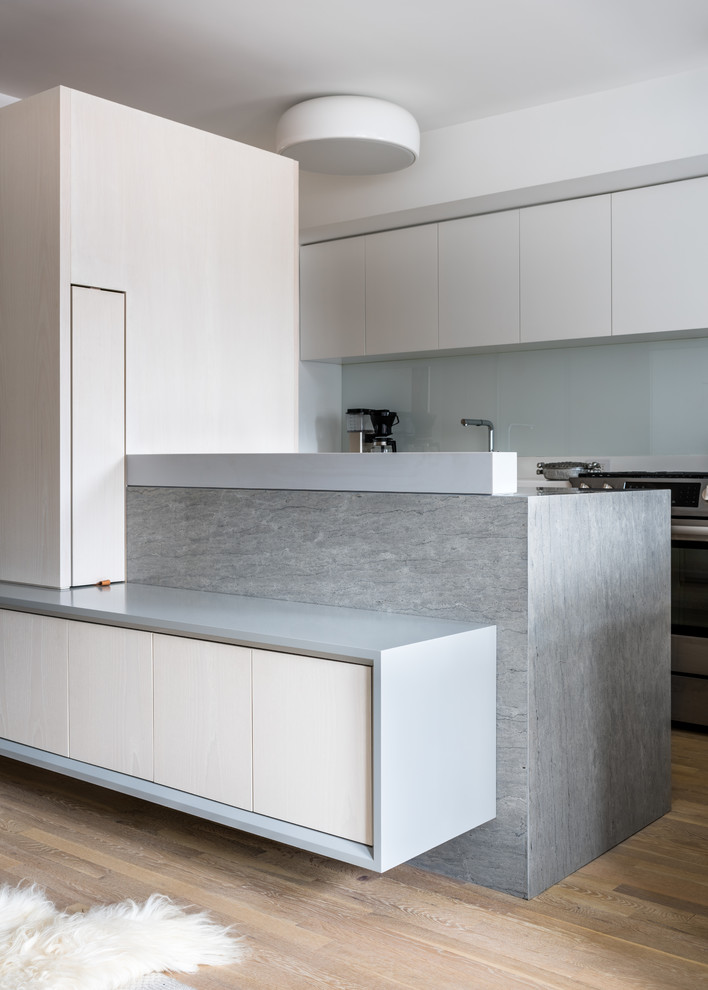 Small contemporary galley kitchen in New York with flat-panel cabinets, white cabinets, glass sheet splashback, light hardwood flooring, a breakfast bar, beige floors, grey worktops, white splashback and stainless steel appliances.