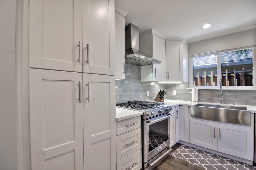 This is an example of a medium sized traditional u-shaped kitchen/diner in San Francisco with a belfast sink, shaker cabinets, white cabinets, engineered stone countertops, grey splashback, glass sheet splashback, stainless steel appliances, medium hardwood flooring, an island, brown floors and white worktops.