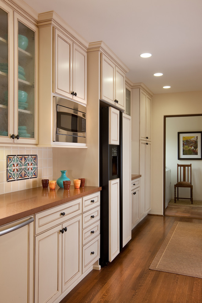 Example of a tuscan medium tone wood floor kitchen design in San Diego with a double-bowl sink, raised-panel cabinets, white cabinets, beige backsplash, ceramic backsplash, stainless steel appliances and no island