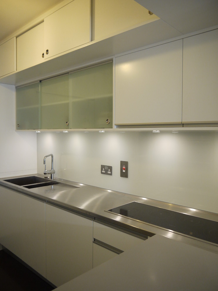 Modern inredning av ett mellanstort kök, med en integrerad diskho, luckor med glaspanel, vita skåp, bänkskiva i rostfritt stål, vitt stänkskydd, glaspanel som stänkskydd, integrerade vitvaror och vinylgolv