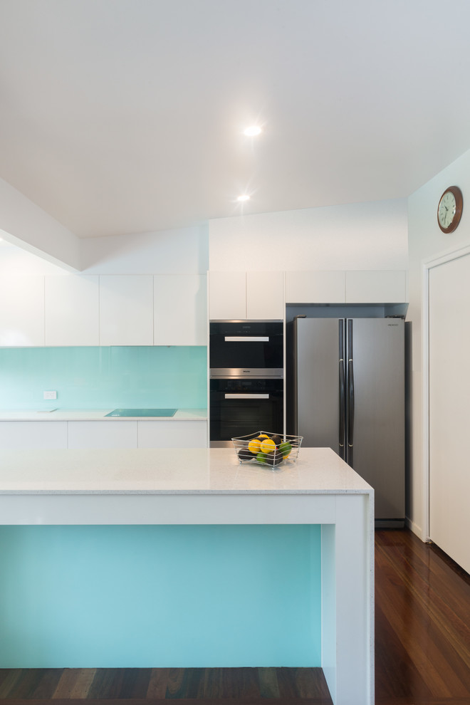 Große Maritime Wohnküche in L-Form mit Einbauwaschbecken, profilierten Schrankfronten, weißen Schränken, Quarzwerkstein-Arbeitsplatte, Küchenrückwand in Blau, Glasrückwand, Küchengeräten aus Edelstahl, braunem Holzboden und Kücheninsel in Brisbane