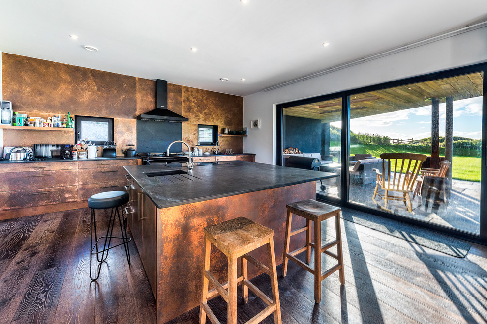 Design ideas for a large urban single-wall kitchen in Cornwall with a submerged sink, flat-panel cabinets, distressed cabinets, copper worktops, black splashback, integrated appliances, dark hardwood flooring, a breakfast bar and brown floors.