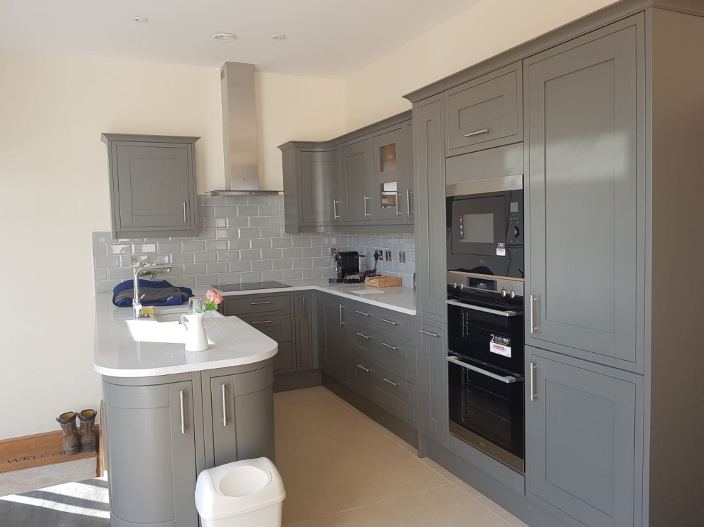 Small traditional u-shaped open plan kitchen in Other with a belfast sink, recessed-panel cabinets, grey cabinets, laminate countertops, grey splashback, ceramic splashback, integrated appliances, porcelain flooring, a breakfast bar, beige floors and white worktops.