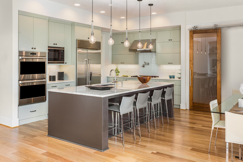 Inspiration for a large classic l-shaped kitchen/diner in Los Angeles with flat-panel cabinets, green cabinets, white splashback, ceramic splashback, stainless steel appliances, an island, a submerged sink, medium hardwood flooring and brown floors.
