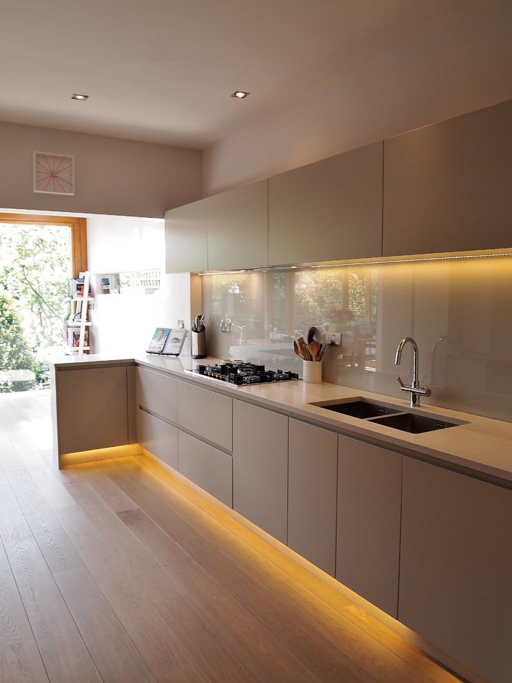 This is an example of a small contemporary galley kitchen/diner in London with a submerged sink, flat-panel cabinets, grey cabinets, engineered stone countertops, green splashback, glass sheet splashback, stainless steel appliances, light hardwood flooring and a breakfast bar.