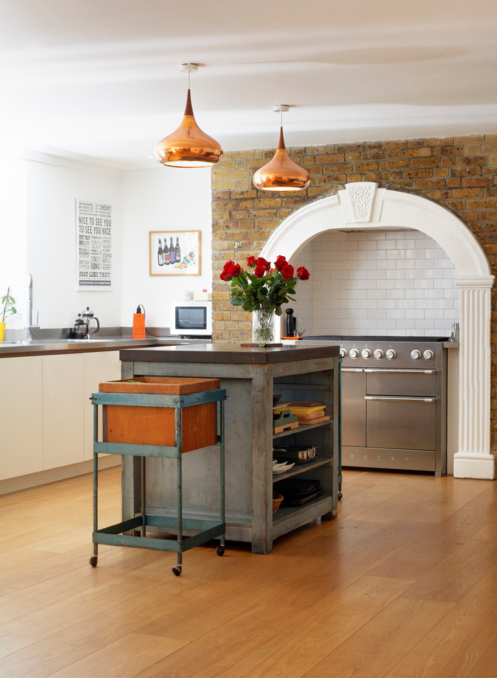 Photo of a victorian kitchen in London with flat-panel cabinets, white cabinets, stainless steel worktops, white splashback, metro tiled splashback, stainless steel appliances, medium hardwood flooring and an island.