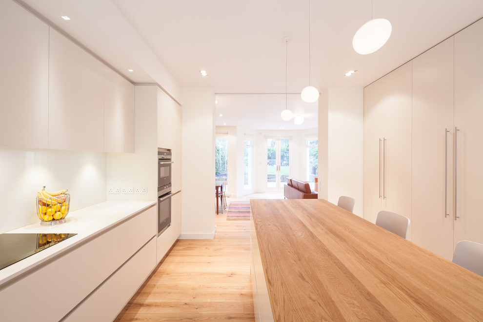 Design ideas for a large contemporary single-wall open plan kitchen in London with a double-bowl sink, flat-panel cabinets, white cabinets, quartz worktops, white splashback, glass sheet splashback, stainless steel appliances, light hardwood flooring, an island, beige floors and white worktops.