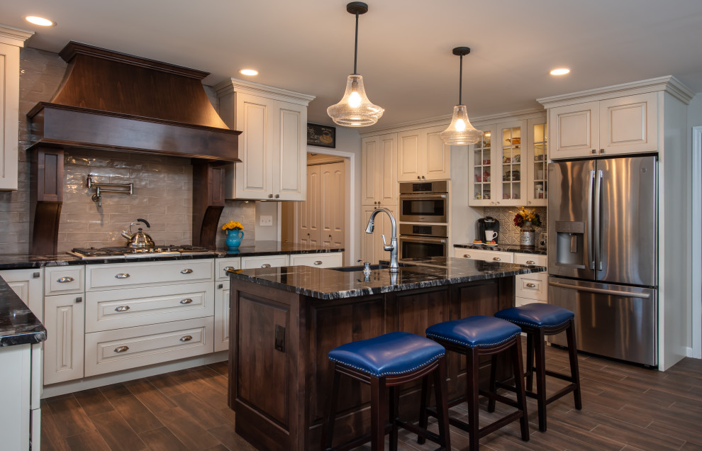 Large transitional l-shaped porcelain tile and brown floor eat-in kitchen photo in Other with a farmhouse sink, raised-panel cabinets, distressed cabinets, granite countertops, beige backsplash, glass tile backsplash, stainless steel appliances, an island and black countertops