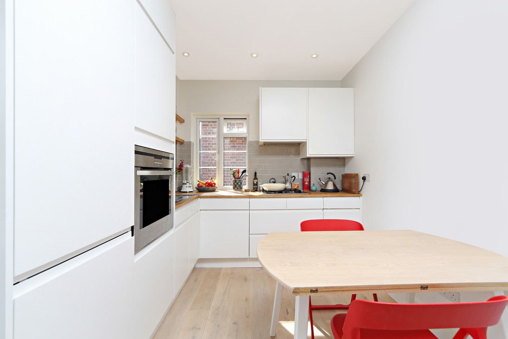 Foto på ett litet minimalistiskt kök, med släta luckor, vita skåp, träbänkskiva, grått stänkskydd, stänkskydd i tunnelbanekakel, integrerade vitvaror och ljust trägolv
