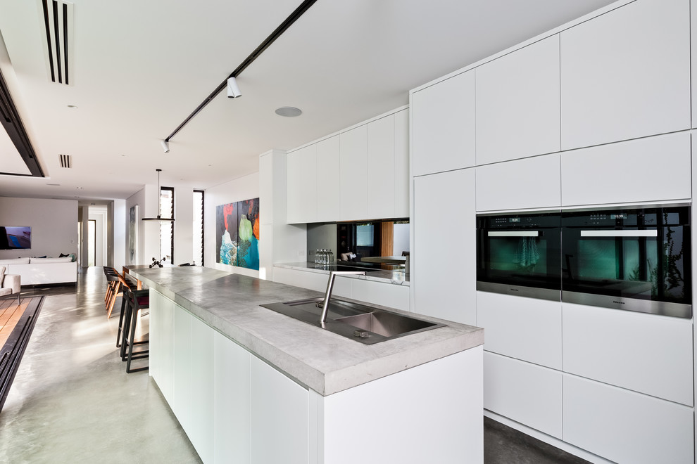 Пример оригинального дизайна: кухня-гостиная в современном стиле с двойной мойкой, плоскими фасадами, белыми фасадами, столешницей из бетона, зеркальным фартуком, черной техникой, бетонным полом, островом и серым полом