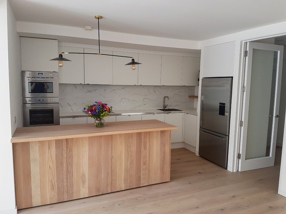 Photo of a small contemporary galley open plan kitchen in London with an integrated sink, flat-panel cabinets, white cabinets, wood worktops, grey splashback, ceramic splashback, stainless steel appliances, light hardwood flooring, a breakfast bar, beige floors and beige worktops.