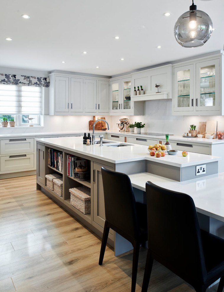 This is an example of a large contemporary u-shaped kitchen/diner in Wiltshire with a double-bowl sink, shaker cabinets, quartz worktops, white splashback, stone slab splashback, black appliances, light hardwood flooring, an island, brown floors and white worktops.