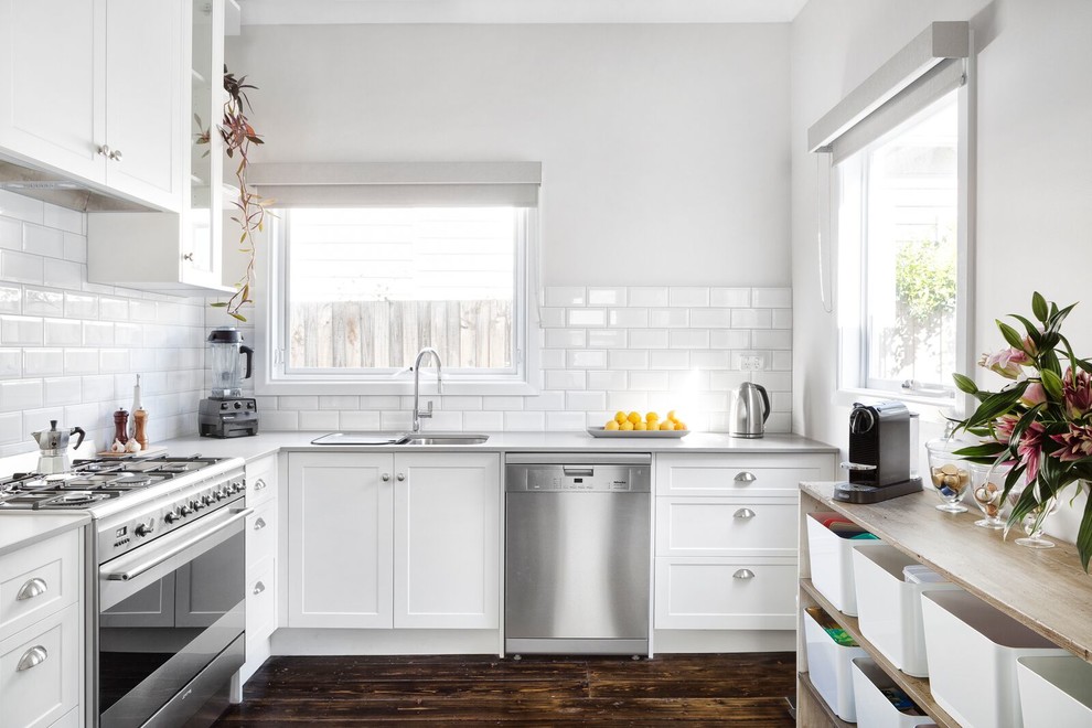 Foto på ett vintage grå l-kök, med en dubbel diskho, skåp i shakerstil, vita skåp, vitt stänkskydd, stänkskydd i tunnelbanekakel, rostfria vitvaror, mörkt trägolv och brunt golv