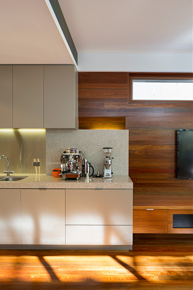This is an example of a contemporary single-wall open plan kitchen in Brisbane with a submerged sink, flat-panel cabinets, beige cabinets, granite worktops, metallic splashback, stainless steel appliances, medium hardwood flooring and an island.