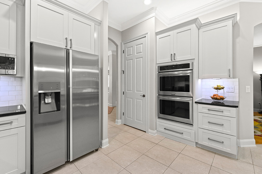 Example of a mid-sized minimalist l-shaped ceramic tile and beige floor eat-in kitchen design in Jacksonville with a farmhouse sink, shaker cabinets, white cabinets, granite countertops, white backsplash, subway tile backsplash, stainless steel appliances, a peninsula and black countertops