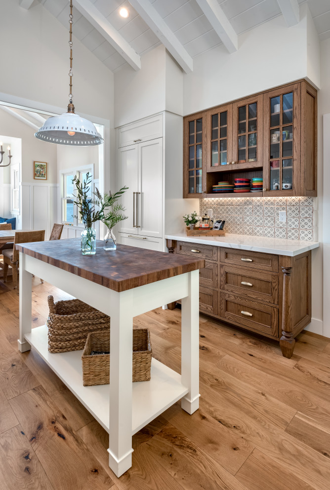 Photo of a medium sized beach style galley kitchen/diner in San Francisco with a belfast sink, raised-panel cabinets, brown cabinets, engineered stone countertops, black splashback, ceramic splashback, integrated appliances, medium hardwood flooring, an island, brown floors and white worktops.