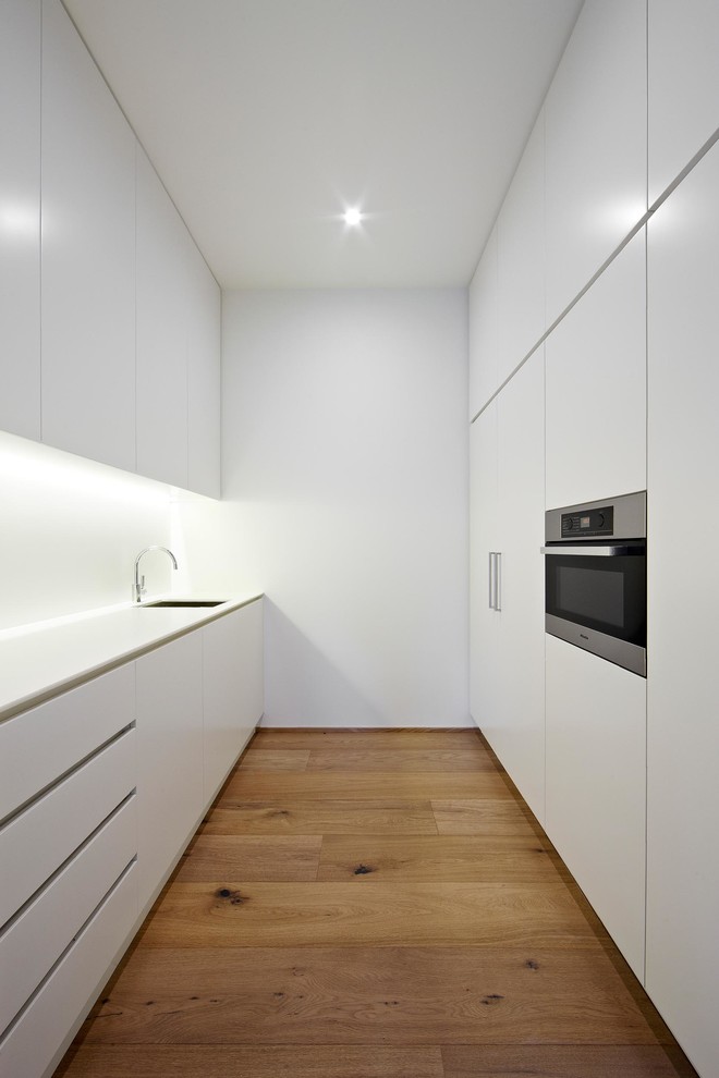 This is an example of a small scandinavian galley enclosed kitchen in Melbourne with flat-panel cabinets, white cabinets, integrated appliances and light hardwood flooring.