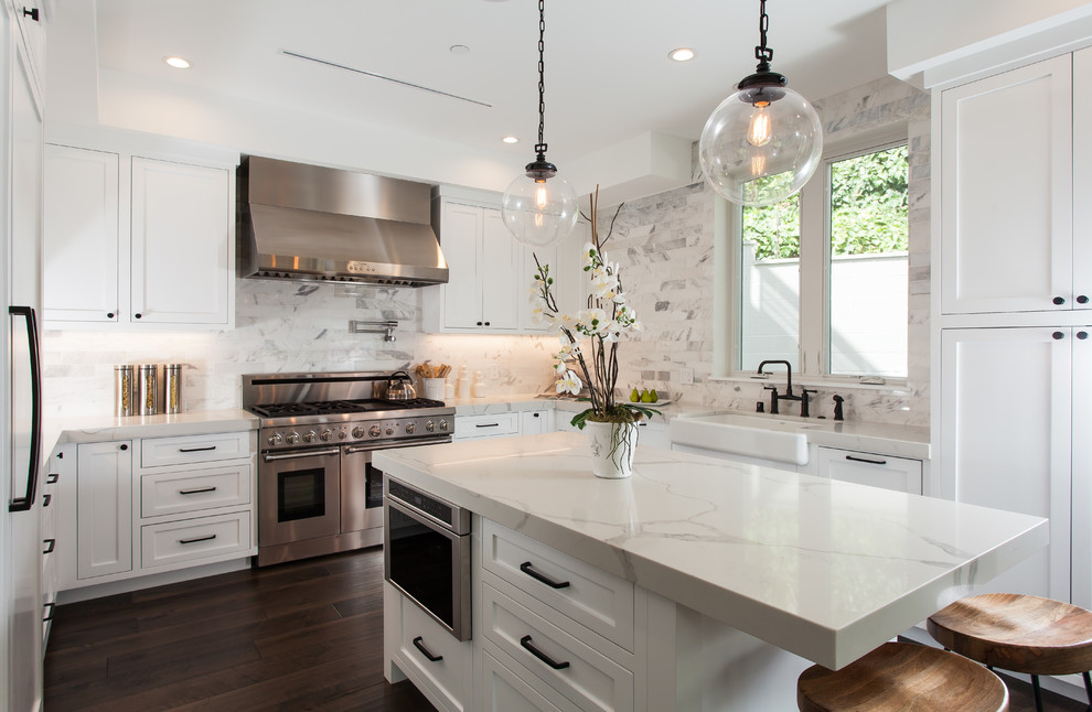 Example of a transitional u-shaped dark wood floor and brown floor enclosed kitchen design in Orange County with a farmhouse sink, shaker cabinets, white cabinets, white backsplash, stainless steel appliances and an island