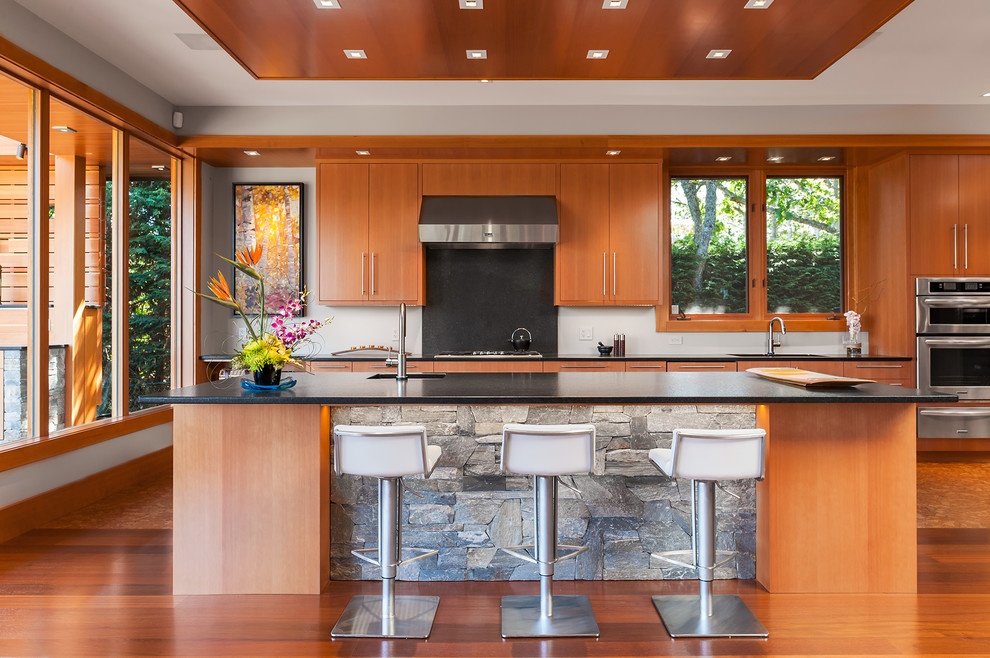 Cette image montre une grande cuisine parallèle design en bois brun avec un placard à porte plane, un électroménager en acier inoxydable, un sol en bois brun, îlot et une crédence noire.