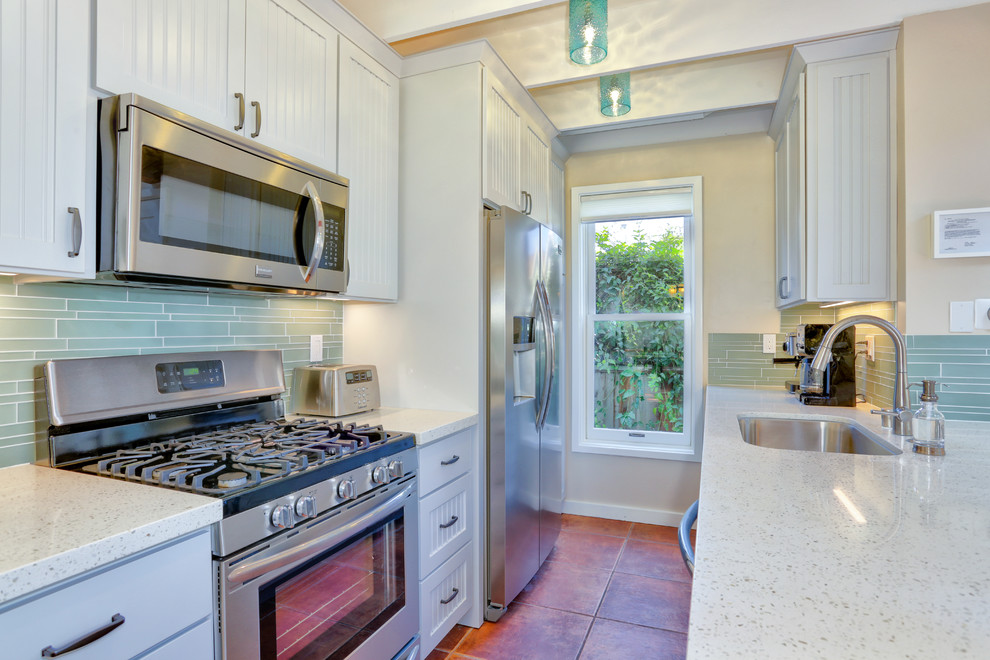 Bild på ett mellanstort maritimt kök, med en enkel diskho, luckor med profilerade fronter, bänkskiva i kvarts, blått stänkskydd, stänkskydd i glaskakel, rostfria vitvaror, vita skåp, klinkergolv i terrakotta, en halv köksö och brunt golv