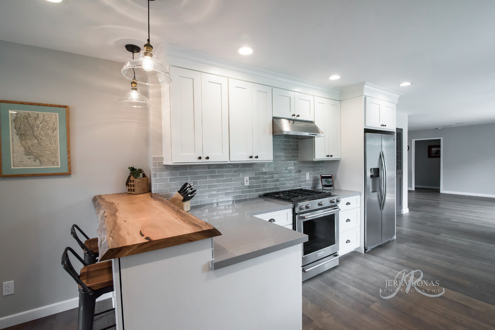 This is an example of a medium sized nautical l-shaped kitchen/diner in San Diego with shaker cabinets, white cabinets, composite countertops, grey splashback, porcelain splashback, stainless steel appliances, dark hardwood flooring, an island and brown floors.