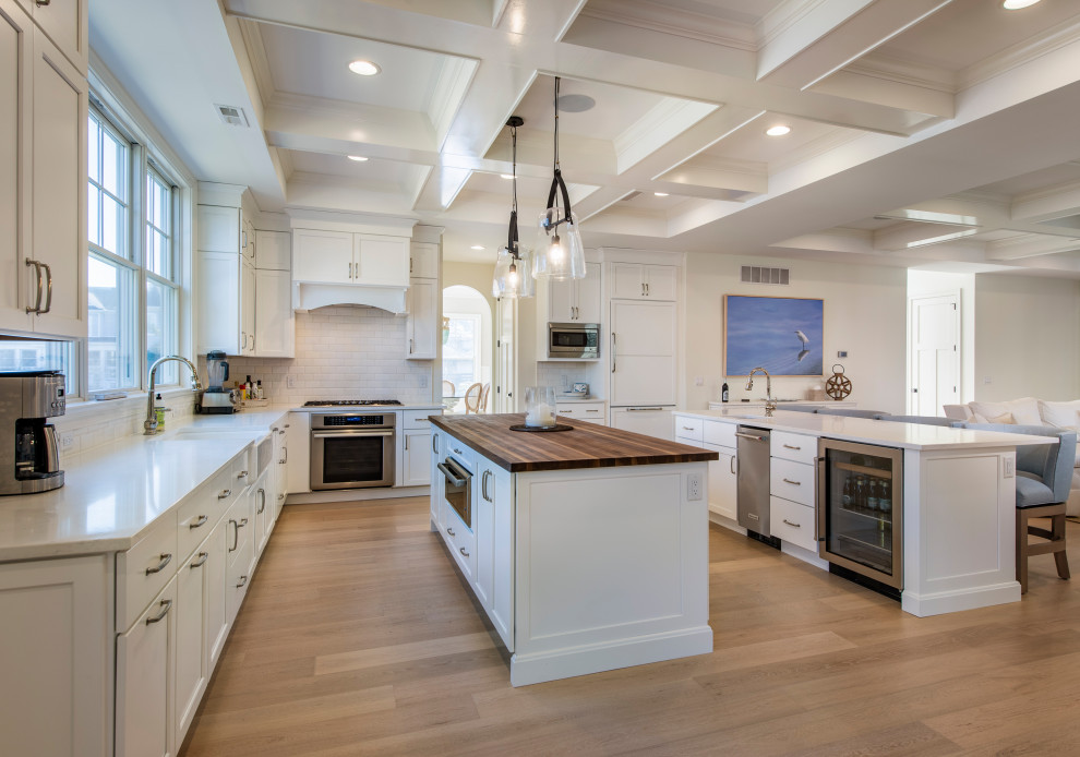 Inspiration for a large coastal l-shaped open plan kitchen in Other with a belfast sink, shaker cabinets, white cabinets, wood worktops, white splashback, marble splashback, medium hardwood flooring, multiple islands, integrated appliances and brown floors.