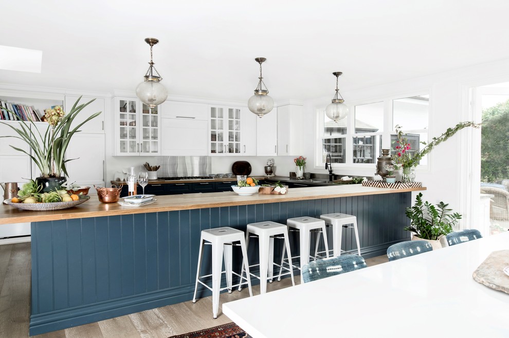 Bild på ett maritimt kök, med en integrerad diskho, skåp i shakerstil, blå skåp, träbänkskiva, vitt stänkskydd, integrerade vitvaror, mellanmörkt trägolv, en halv köksö och grått golv