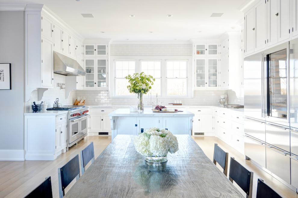 Inspiration for a large beach style u-shaped kitchen/diner in New York with shaker cabinets, white cabinets, white splashback, stainless steel appliances, light hardwood flooring and an island.