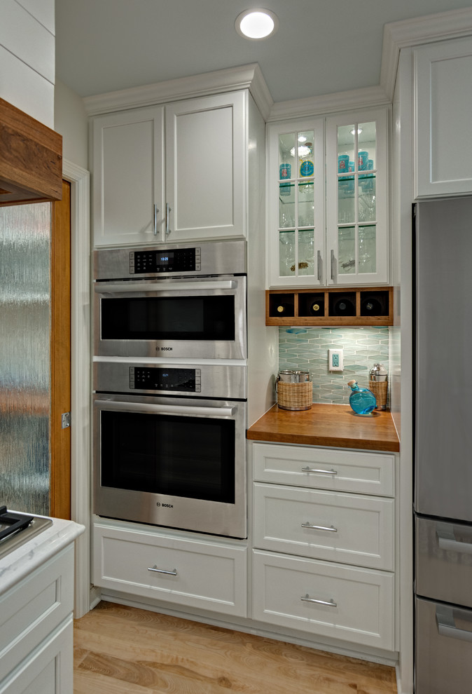 Maritim inredning av ett mellanstort vit vitt kök, med en undermonterad diskho, släta luckor, vita skåp, bänkskiva i kvarts, blått stänkskydd, stänkskydd i glaskakel, rostfria vitvaror, ljust trägolv och en halv köksö