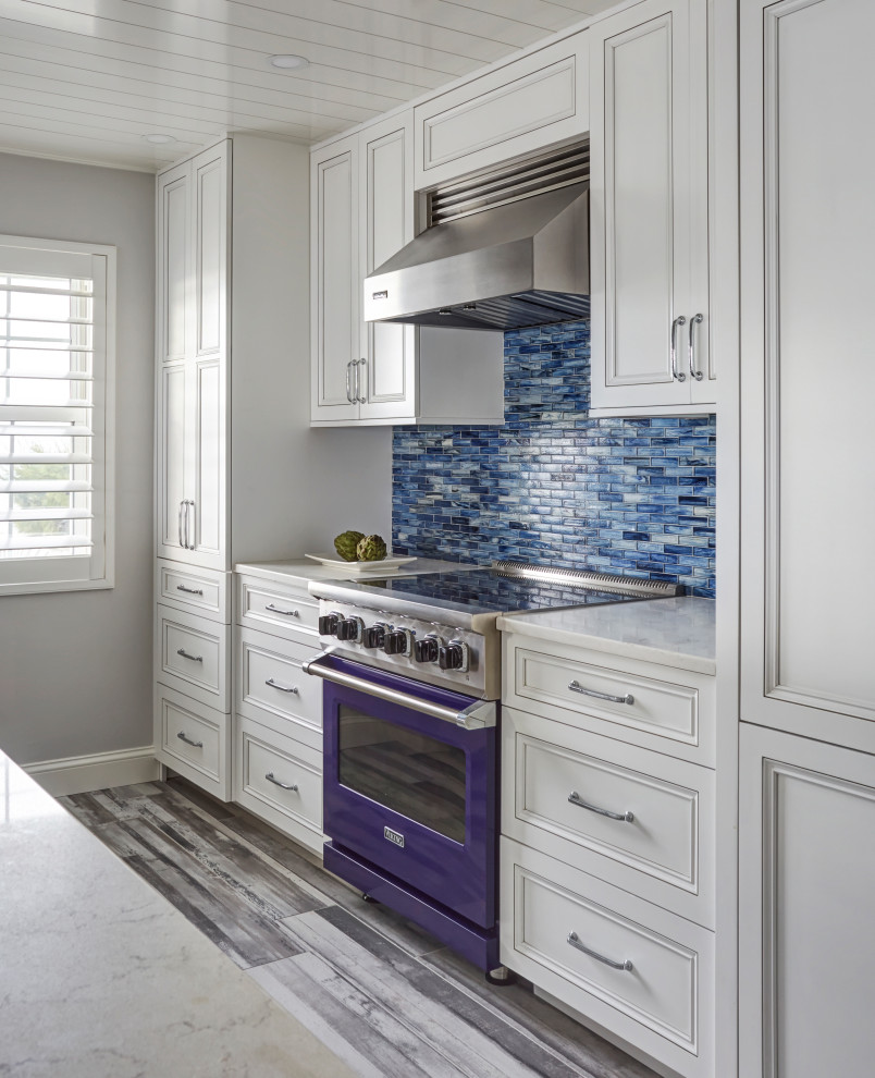 高級なビーチスタイルのおしゃれなキッチン (アンダーカウンターシンク、落し込みパネル扉のキャビネット、白いキャビネット、クオーツストーンカウンター、青いキッチンパネル、ガラスタイルのキッチンパネル、パネルと同色の調理設備、磁器タイルの床、グレーの床、白いキッチンカウンター、塗装板張りの天井) の写真