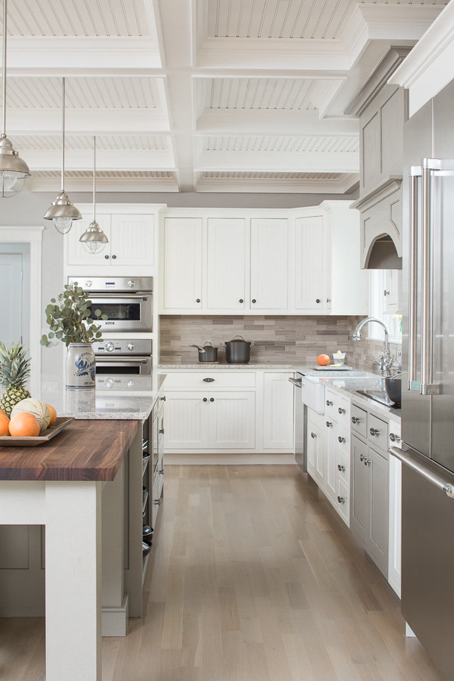 Transitional l-shaped light wood floor kitchen photo in Portland Maine with a farmhouse sink, shaker cabinets, white cabinets, granite countertops, brown backsplash, stainless steel appliances and an island