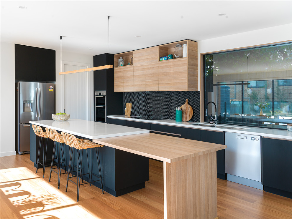 Photo of a large beach style galley open plan kitchen in Other with a submerged sink, black cabinets, engineered stone countertops, black splashback, mosaic tiled splashback, stainless steel appliances, light hardwood flooring, an island, beige floors and white worktops.
