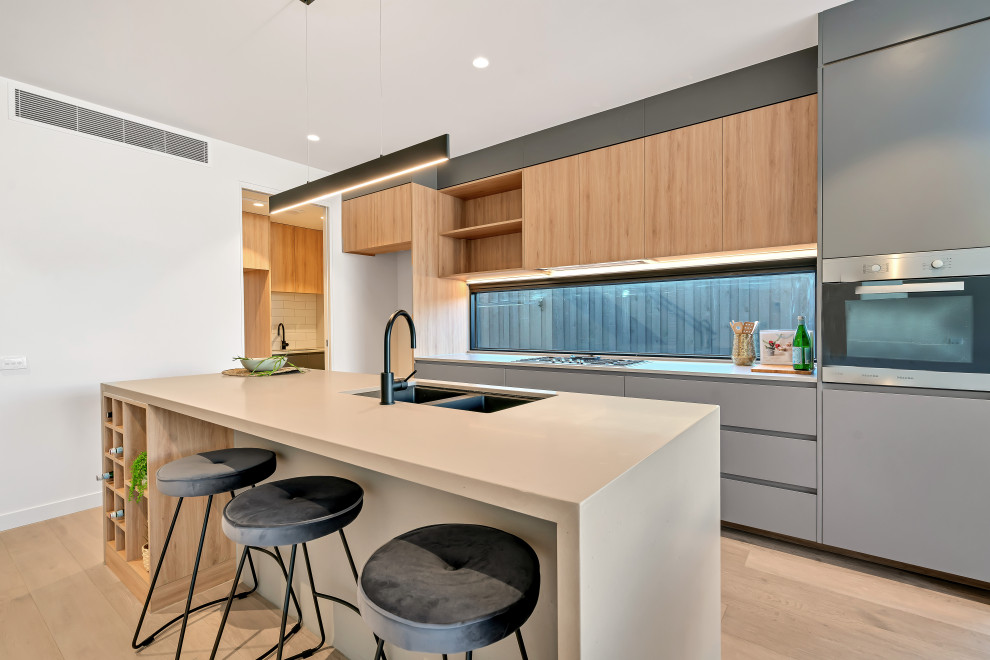 Foto på ett mellanstort funkis grå kök, med en undermonterad diskho, släta luckor, grå skåp, rostfria vitvaror, ljust trägolv, en köksö, bänkskiva i kvarts, fönster som stänkskydd och brunt golv