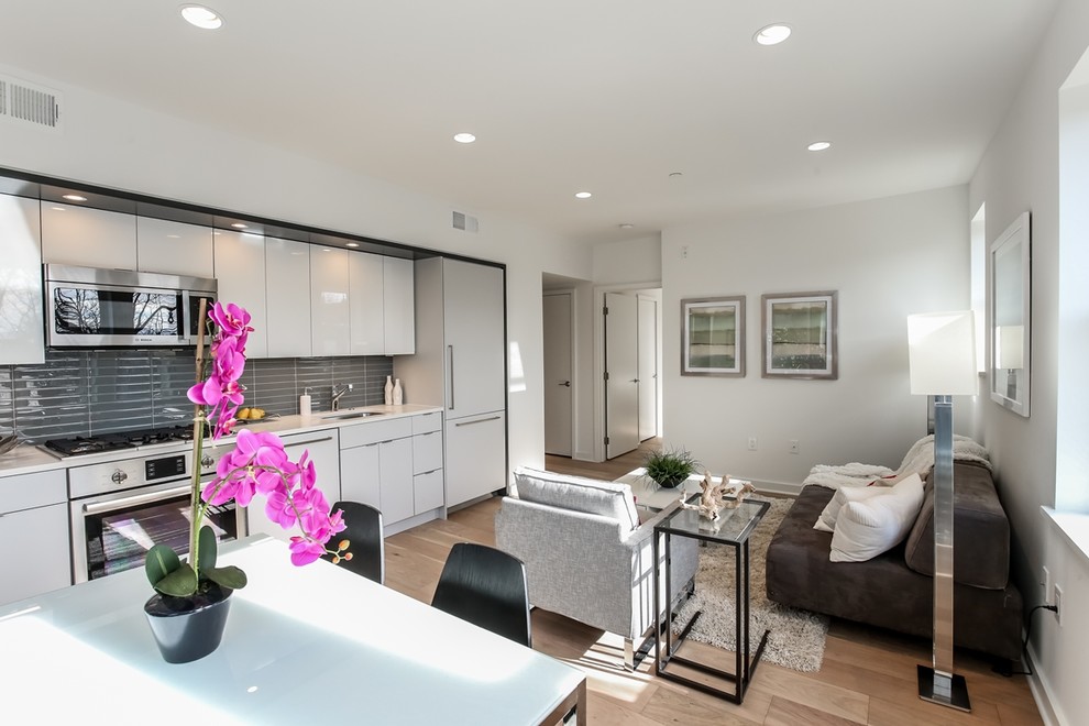 Small modern single-wall kitchen/diner in DC Metro with a built-in sink, flat-panel cabinets, engineered stone countertops, grey splashback, ceramic splashback, integrated appliances, light hardwood flooring, an island and white cabinets.