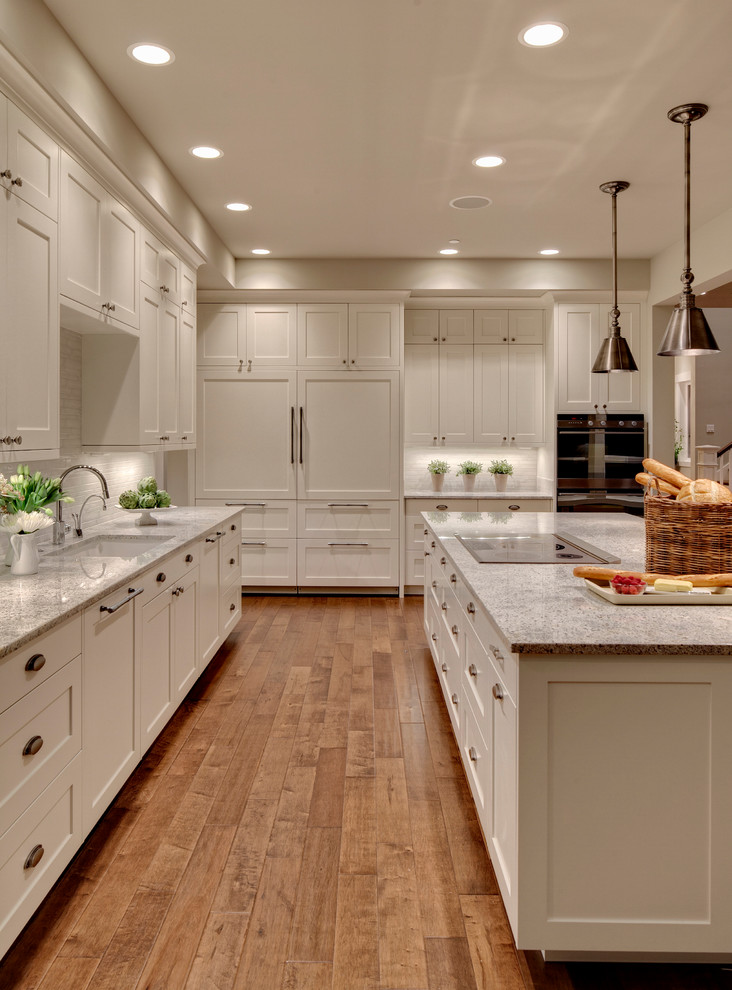 Example of a transitional l-shaped medium tone wood floor open concept kitchen design in Seattle with an undermount sink, recessed-panel cabinets, white cabinets, metallic backsplash, porcelain backsplash, stainless steel appliances and an island