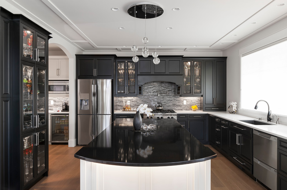 Klassisk inredning av ett stort vit vitt kök, med en undermonterad diskho, luckor med infälld panel, svarta skåp, bänkskiva i kvarts, grått stänkskydd, stänkskydd i glaskakel, rostfria vitvaror, mellanmörkt trägolv, en köksö och brunt golv